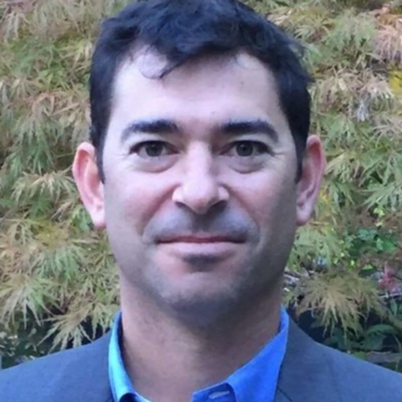 A man in a blue shirt and grey blazer stands before a backdrop of lush, yellowish-green foliage.