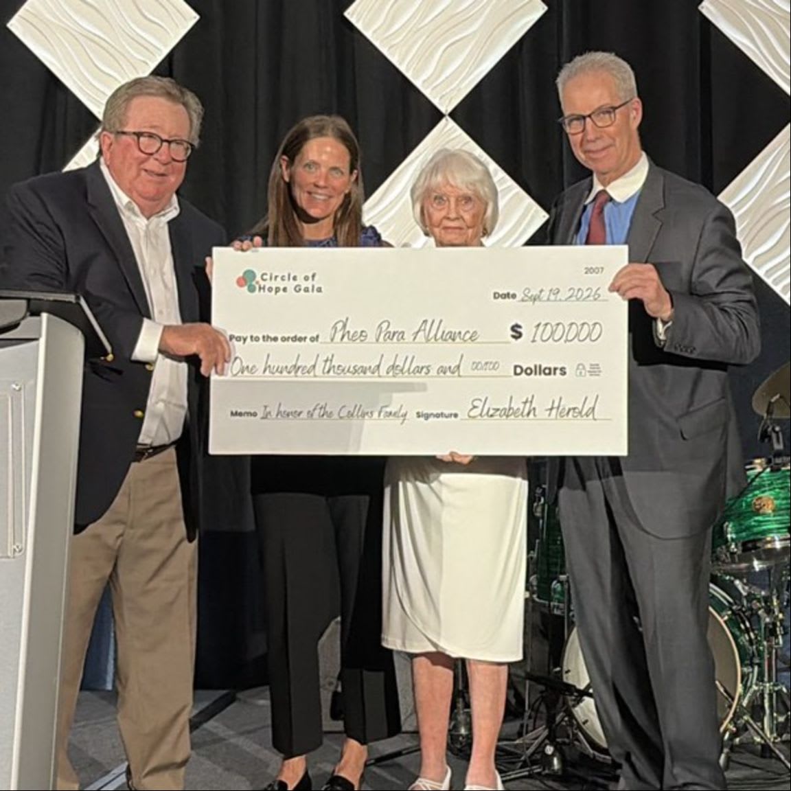Four people stand in the foreground holding a large ceremonial check for $100,000 against a background of black curtains and white diamond patterns.