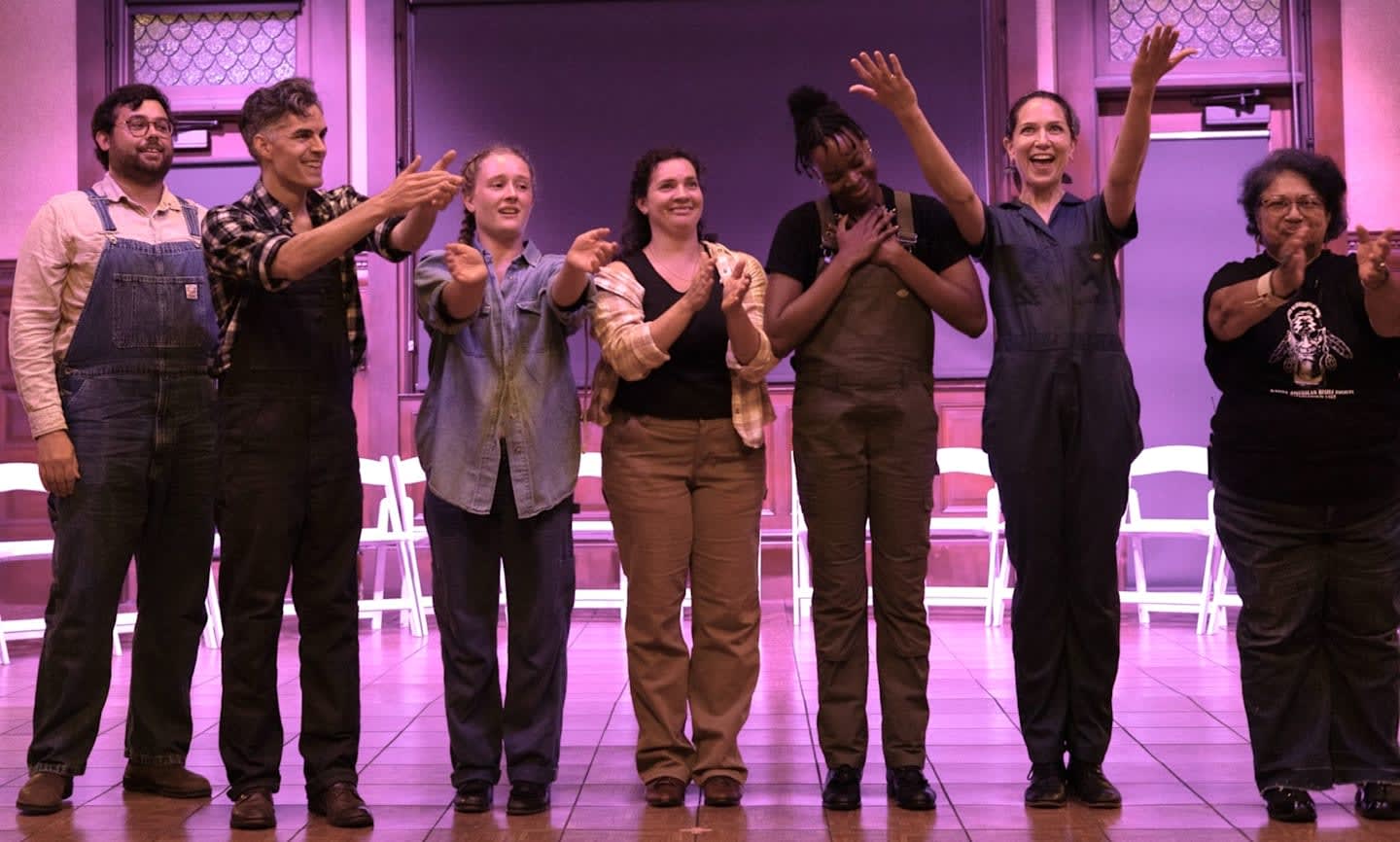 A group of seven people in workwear stand and applaud on a wooden stage in front of a dark background with white chairs.