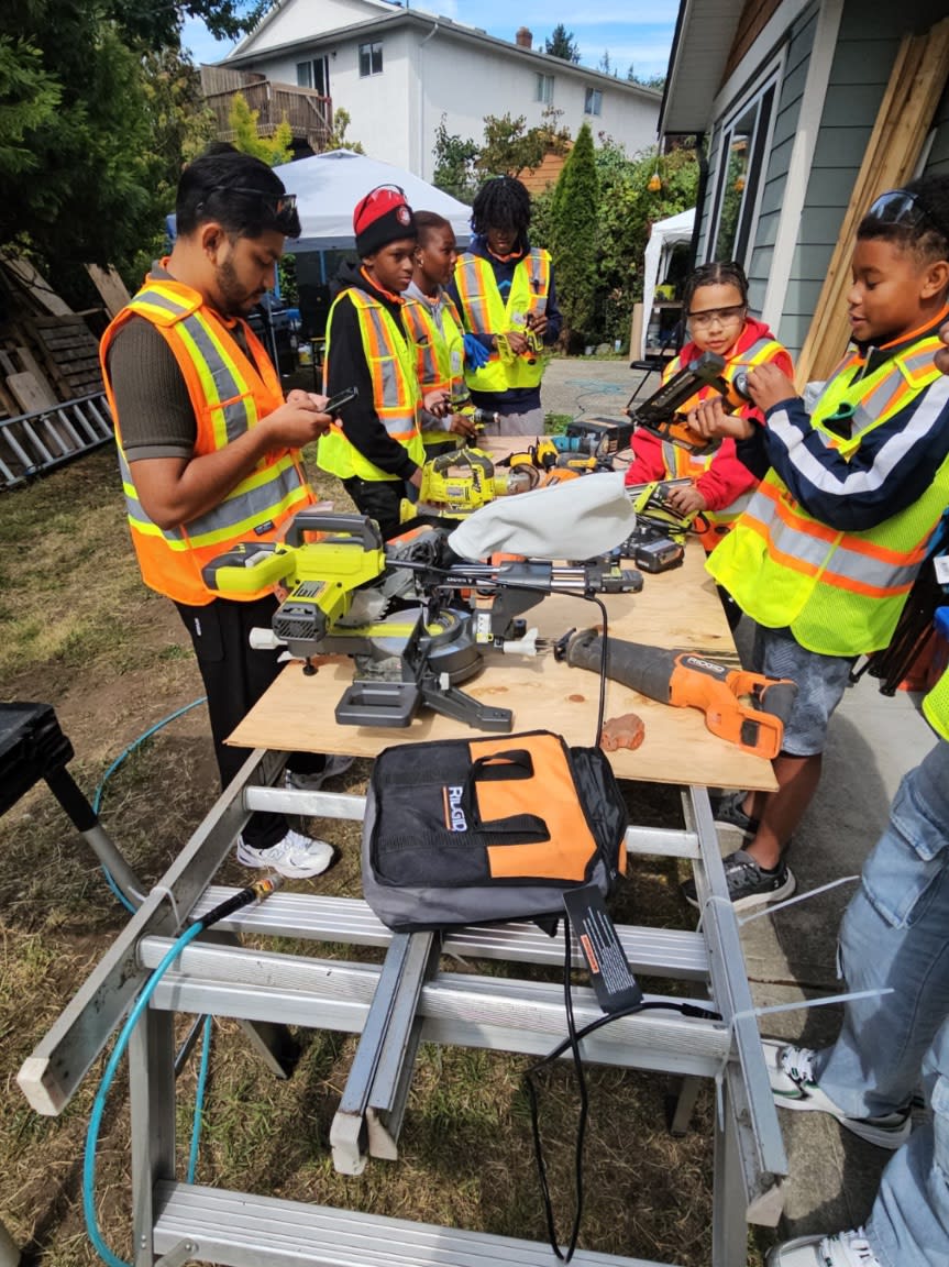 A group of people in high-visibility vests gather around a workbench with power tools in a residential backyard.