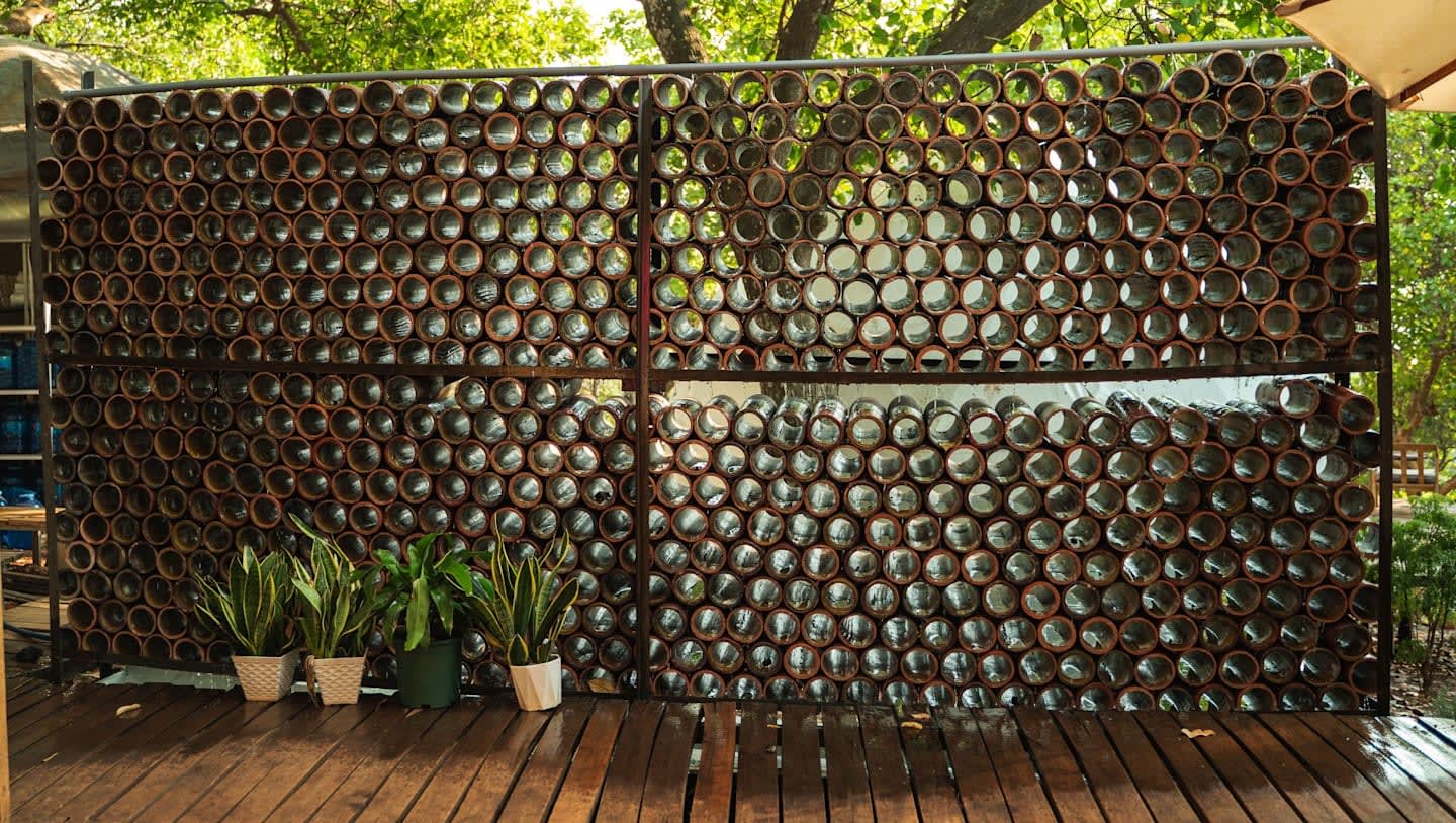A wall made of stacked glass bottles stands on a wooden deck with potted plants in the foreground and lush green trees in the background.