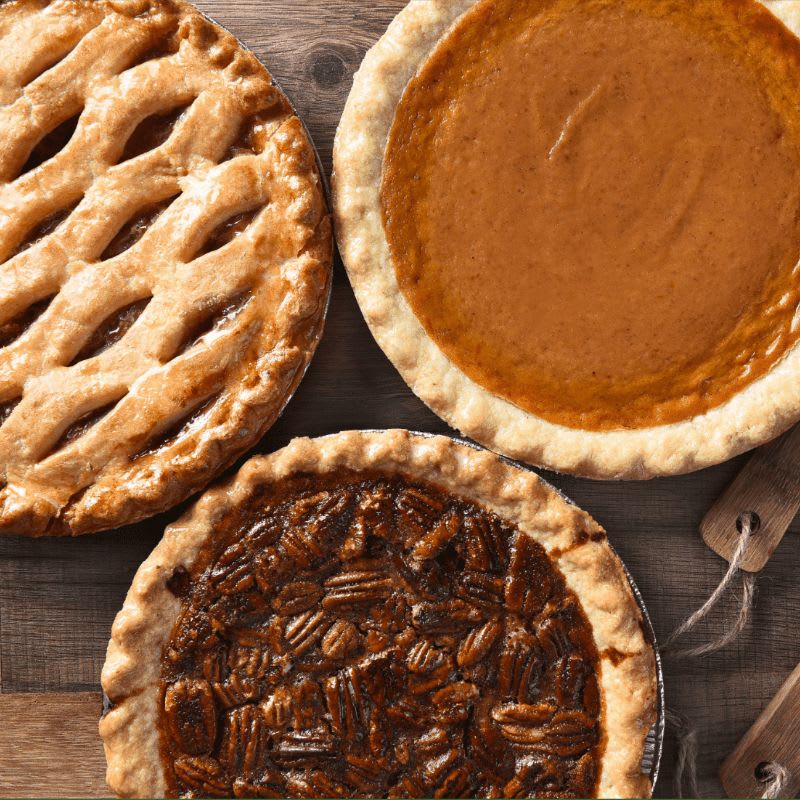Three different pies—lattice-crust, pumpkin, and pecan—rest on a rustic wooden surface.