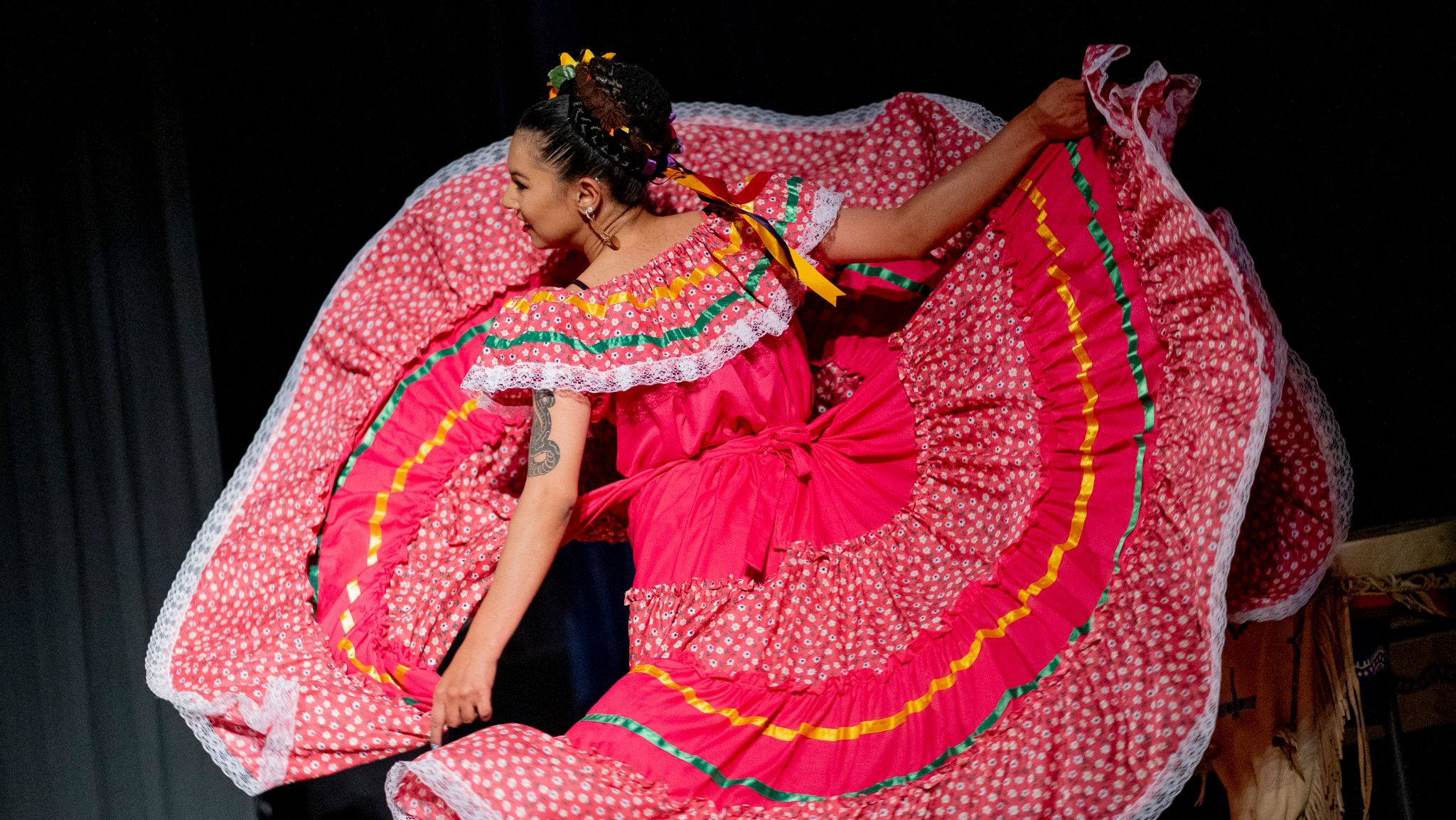 A dancer dressed in a traditional Mexican folkloric dress twirls gracefully on stage. The vibrant pink dress features floral patterns, colorful ruffled stripes, and white lace trim, creating a striking visual as it fans out around her. She has braided hair adorned with colorful ribbons, and her tattooed arm is visible as she extends it gracefully. The background is dark, emphasizing the movement and vivid colors of her attire.