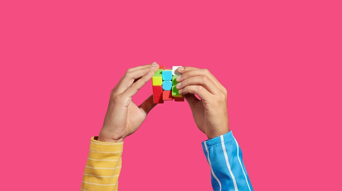 Two hands working to solve a puzzle cube; the hand on the left sits atop a yellow sleeve and the hand on the right is coming out of a blue sleeve.