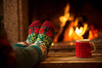 A person wearing thick socks rests before a fire with a warm drink during the holidays.