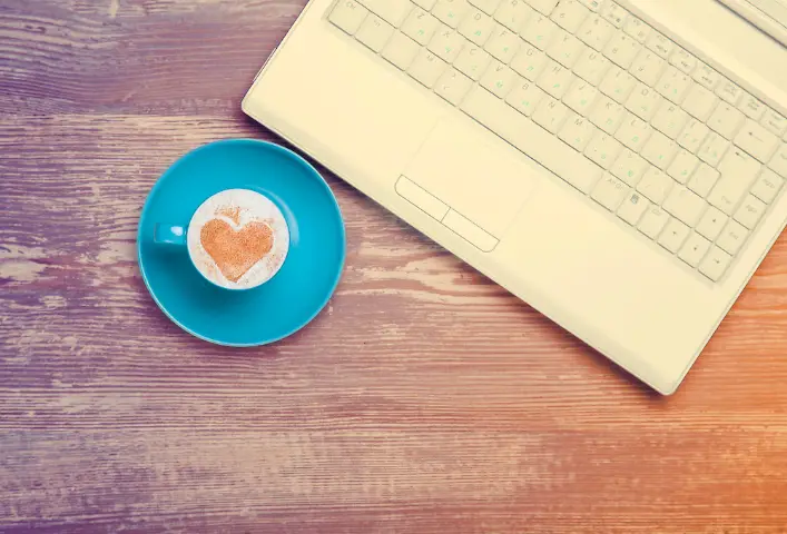 Image of a coffee cup in a saucer near a laptop, delicious coffee with cream and cinnamon sprinkled in the shape of a heart.