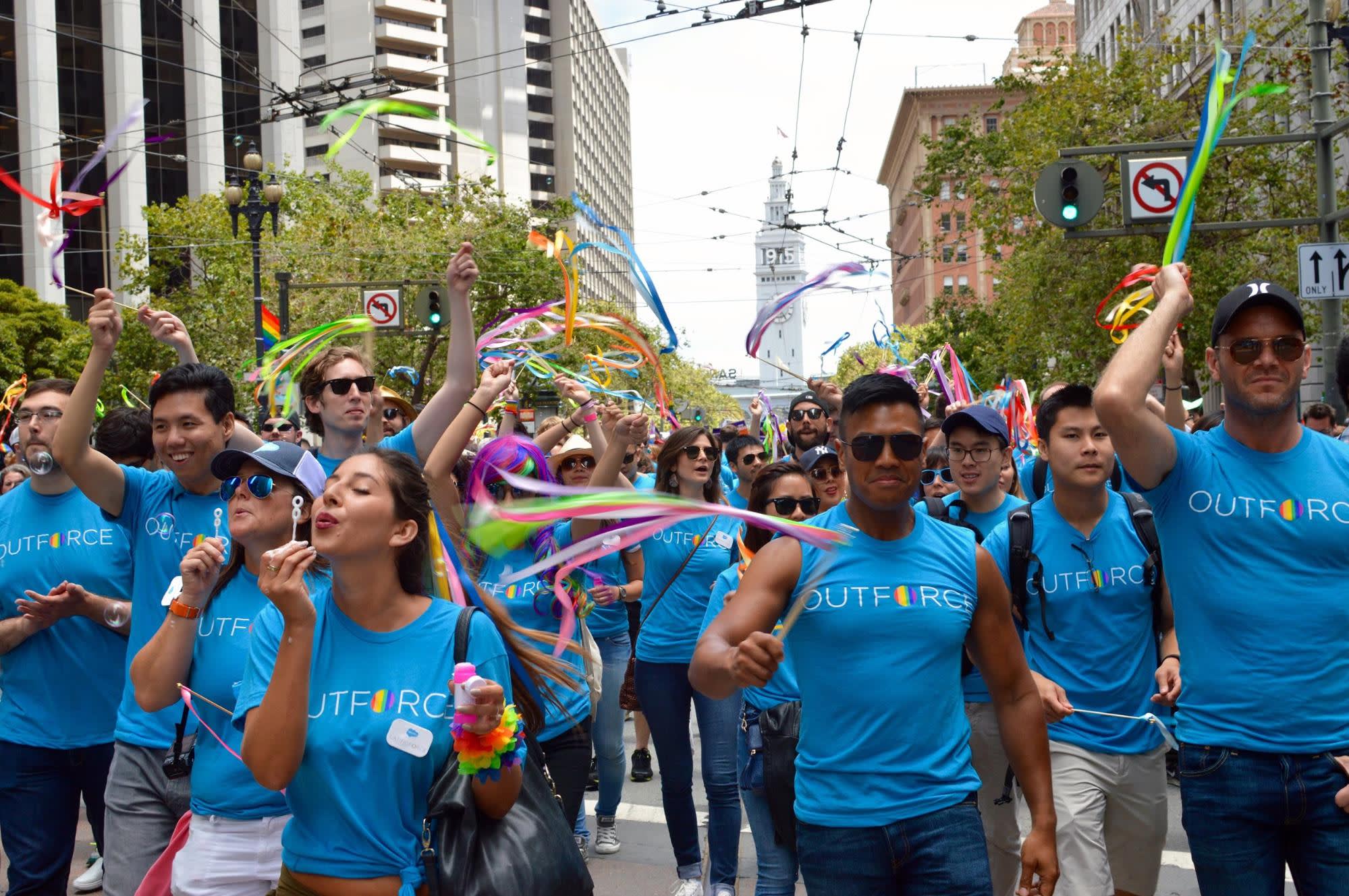 Picture of a Pride parade, with people marching in Outforce tshirts, blowing bubbles, and waving streamers