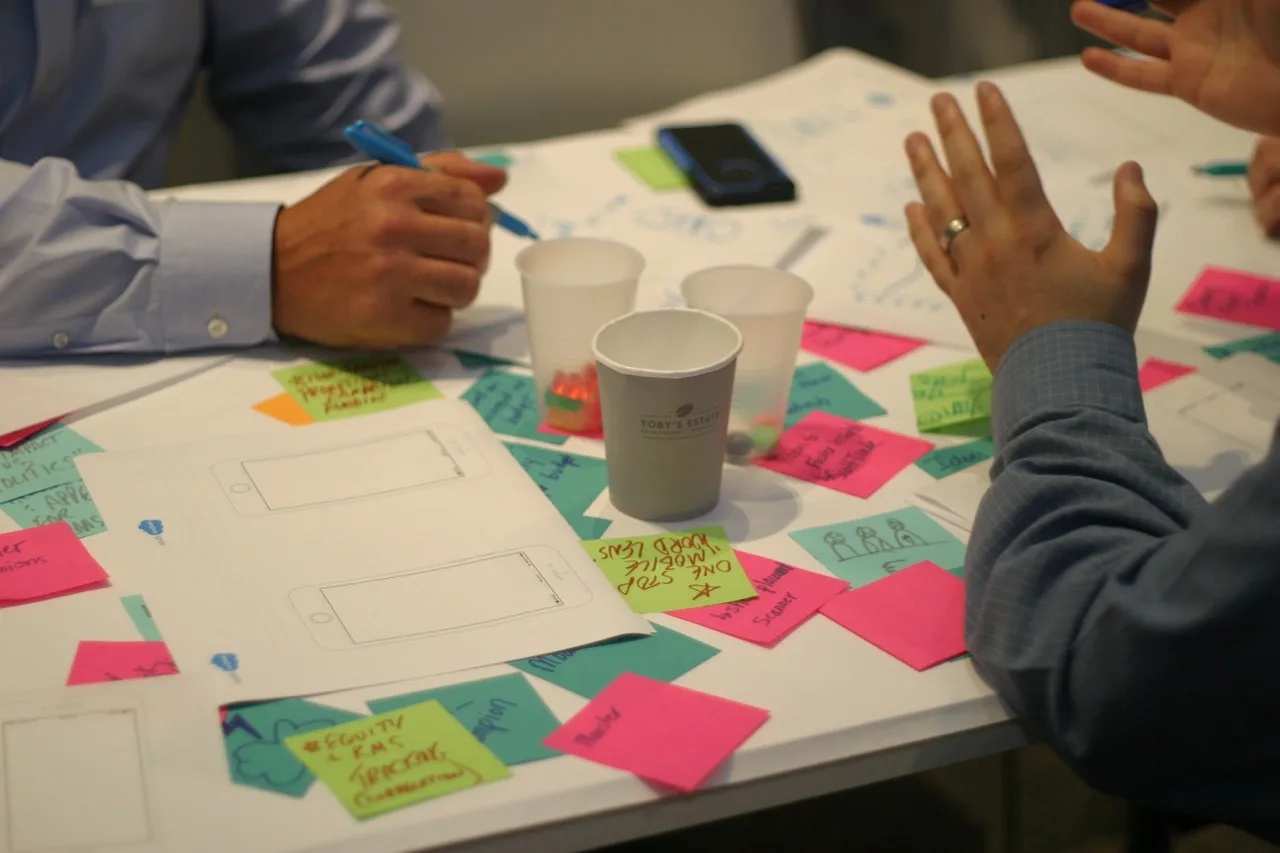 Photo of work table, showing cups, papers, post-its, and hands gesturing.