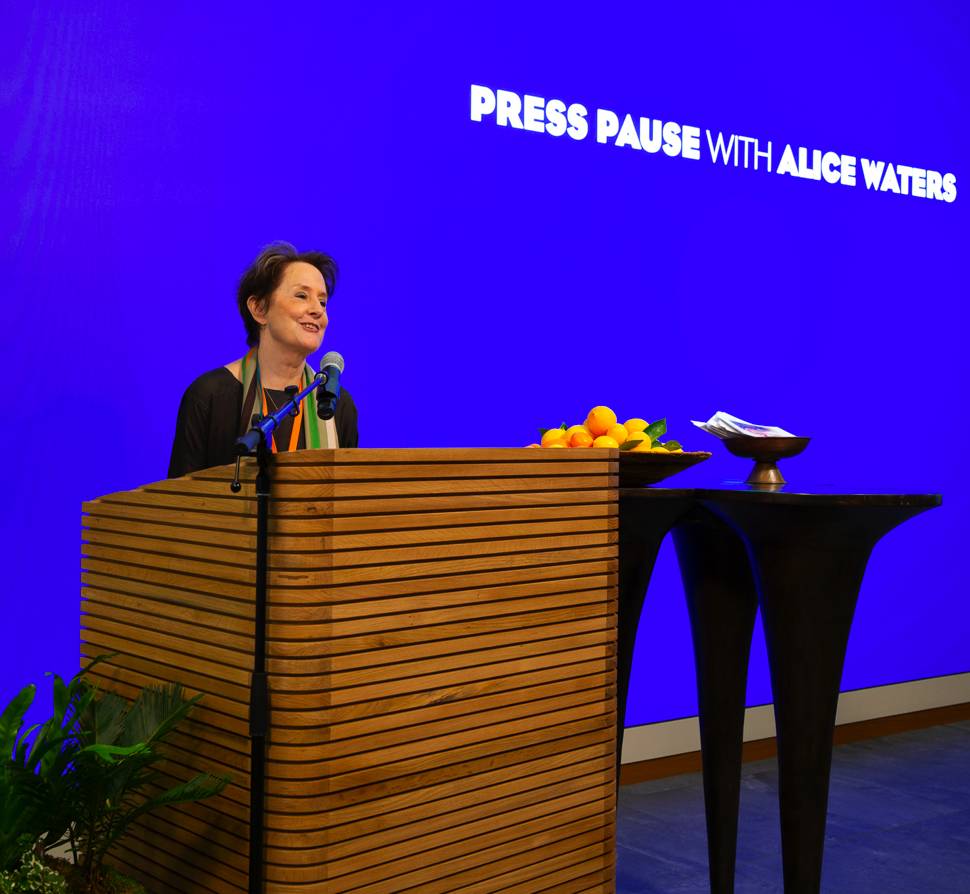 An image of Alice Waters speaking at Salesforce with “Press Pause with Alice Waters” displayed on a screen in the background.