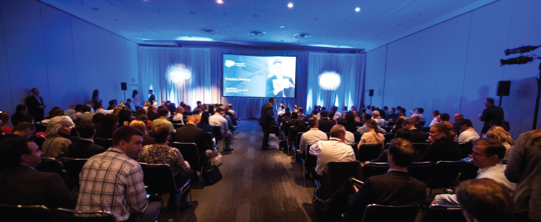 Large, blue-lit presentation room with audience.