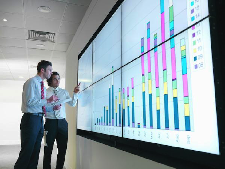 Two men in front of a video screen displaying a chart