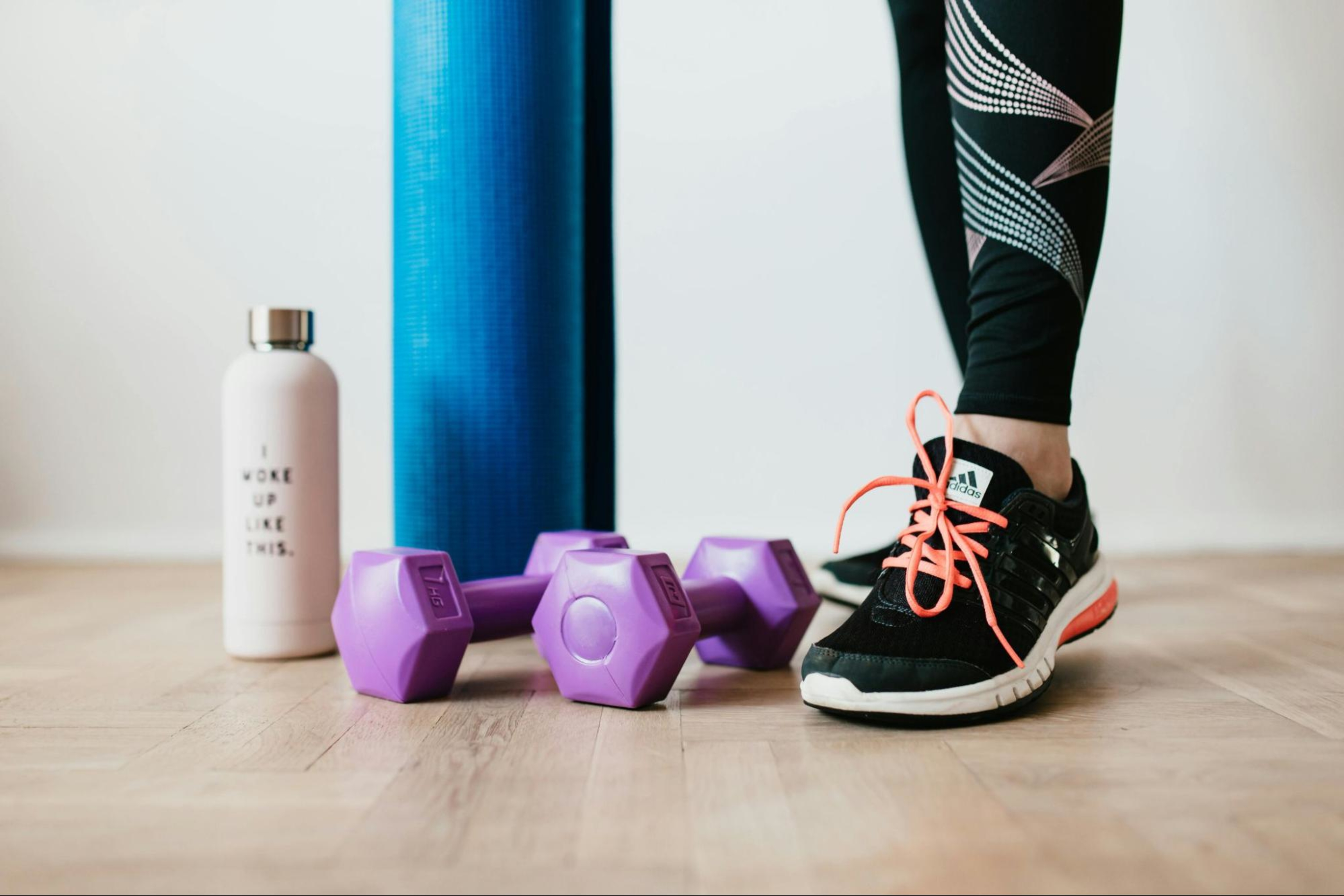 A working professional stands with a yoga mat, water bottle, and dumbbells to do an at-home workout.