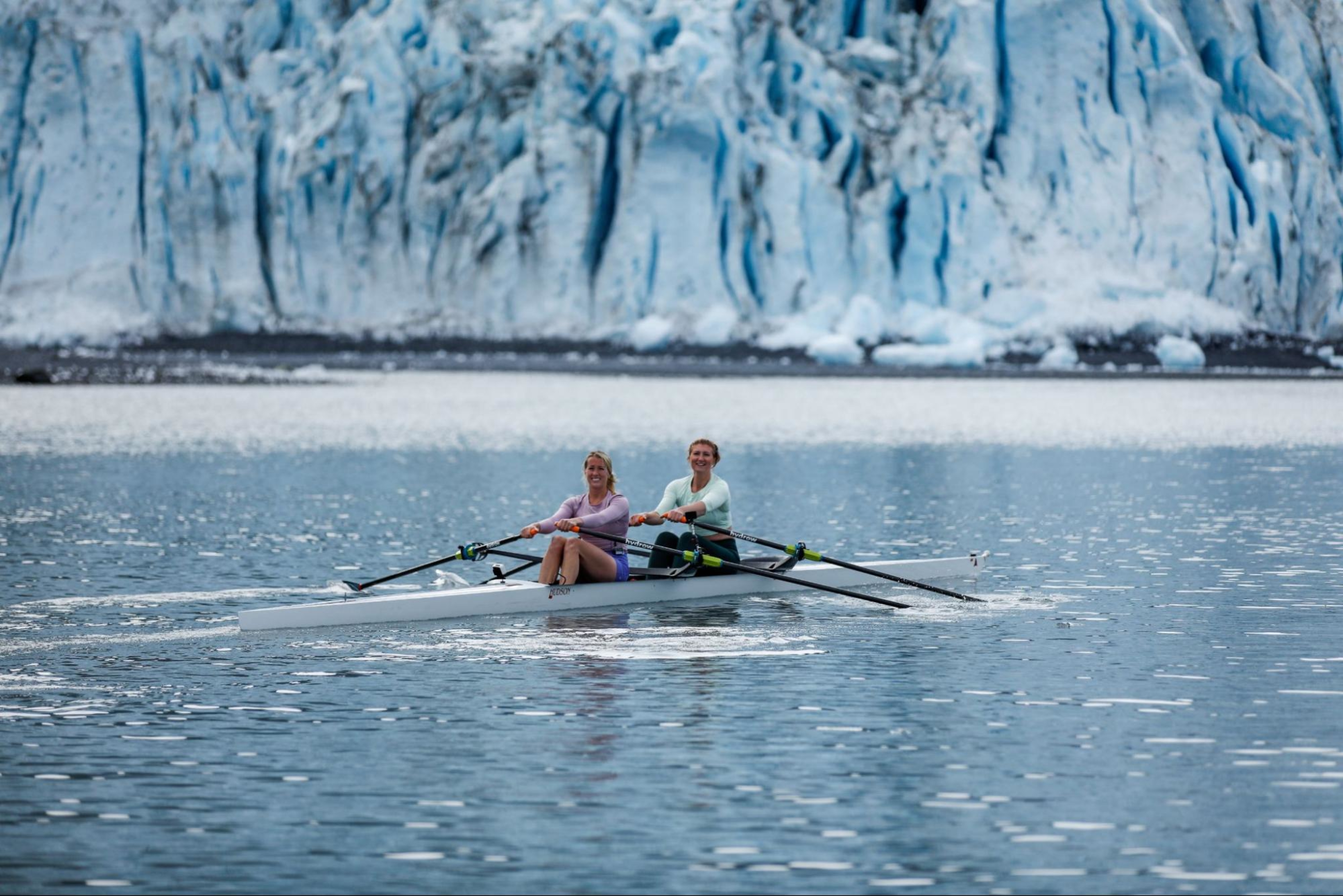 Two Hydrow Athletes lead a rowing workout from the water in Alaska, which is included as part of a Hydrow membership.