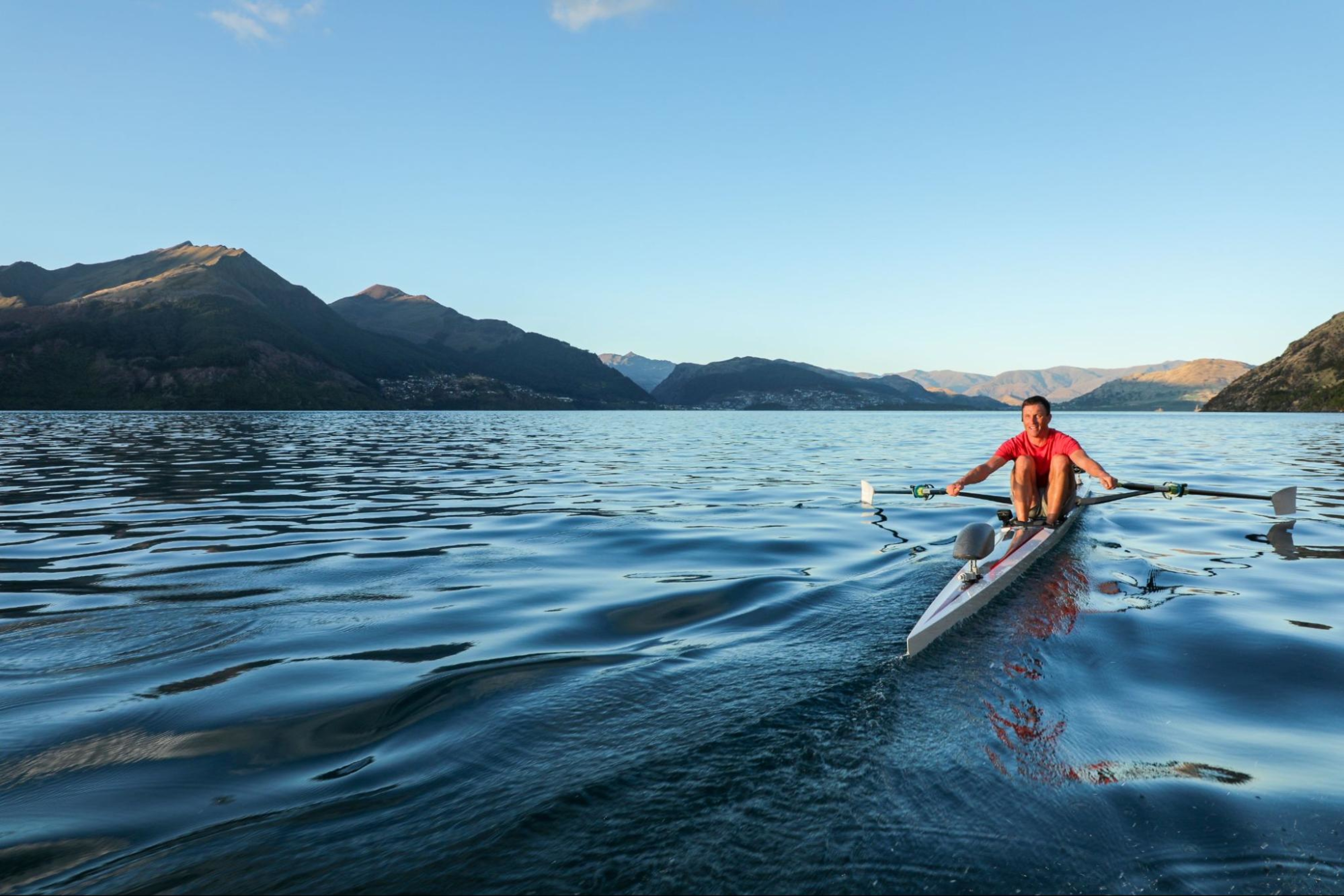 Man does a rowing workout on the water with mountains in the background.