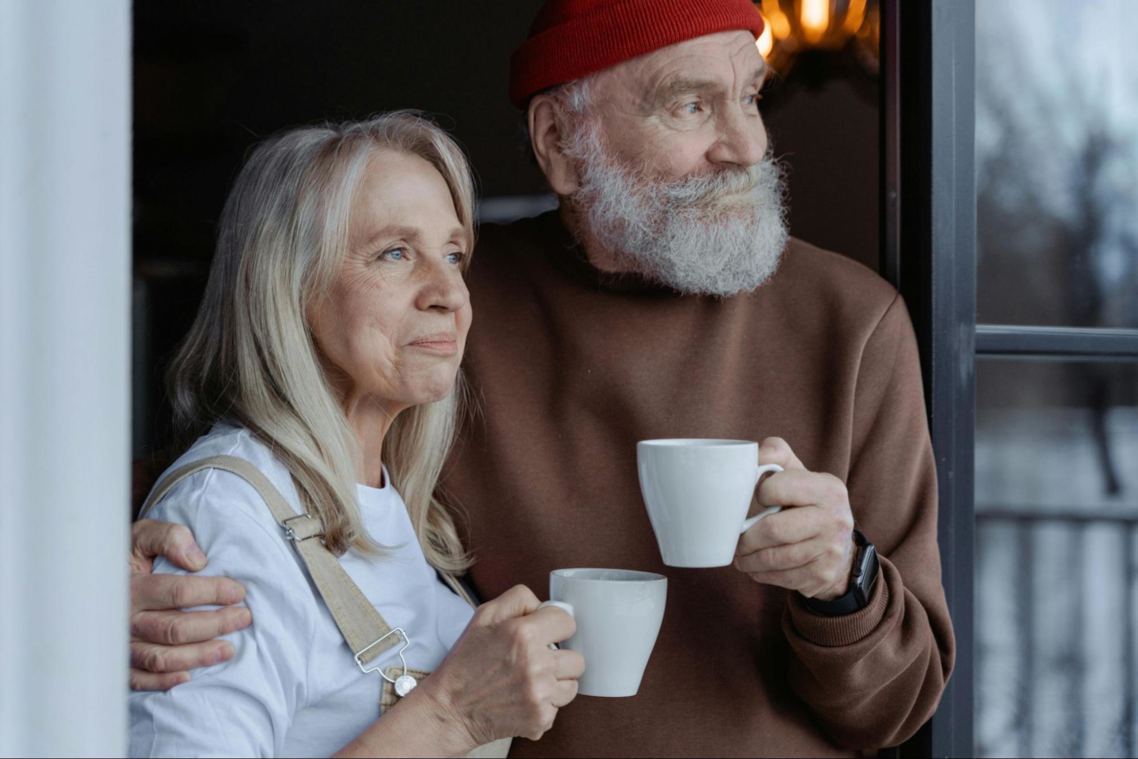 Older couple embraces while looking outside and holding coffee cups.