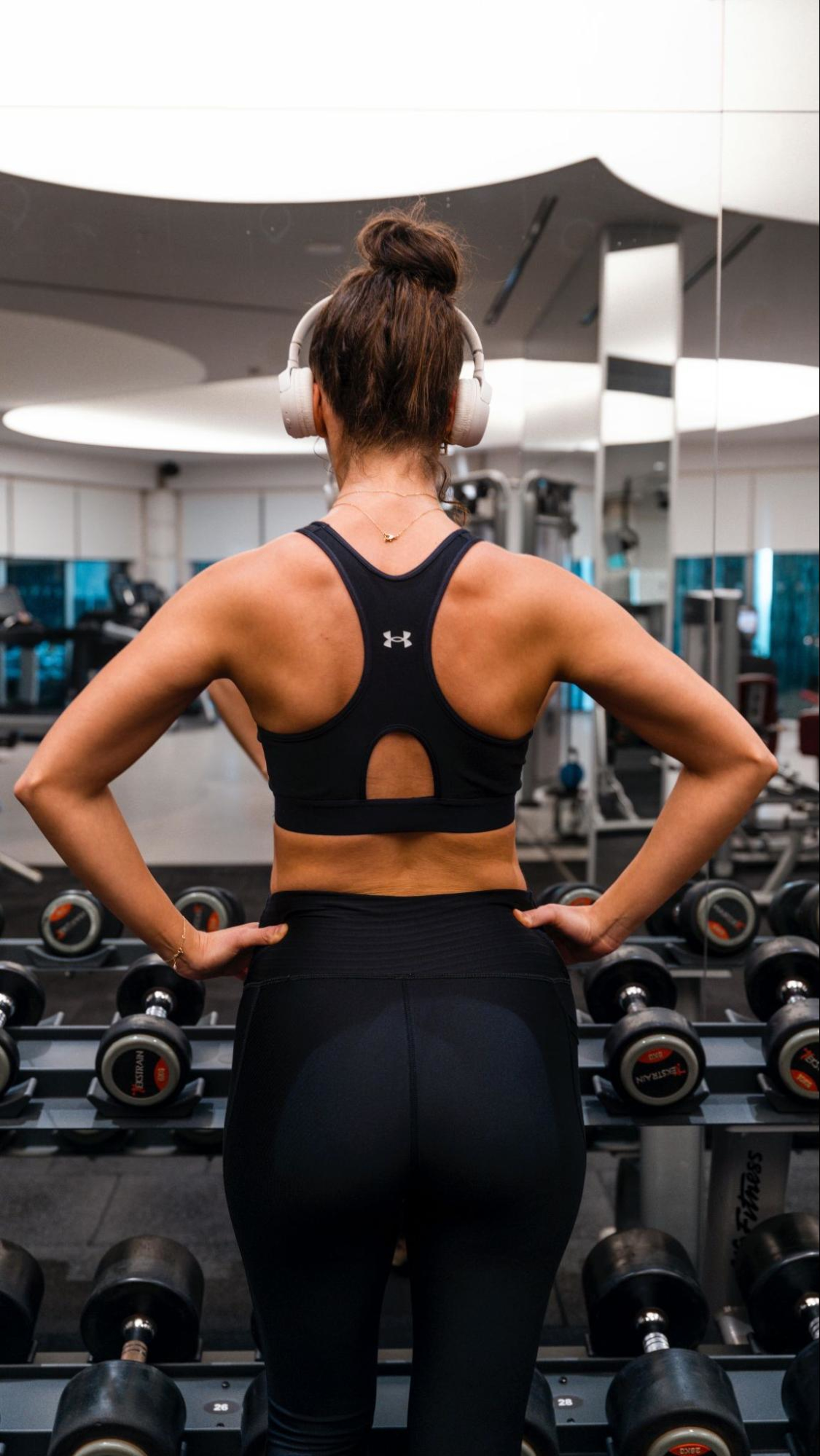 Woman wearing headphones stands in front of a dumbbell rack as she learns how to build muscle mass in glutes.