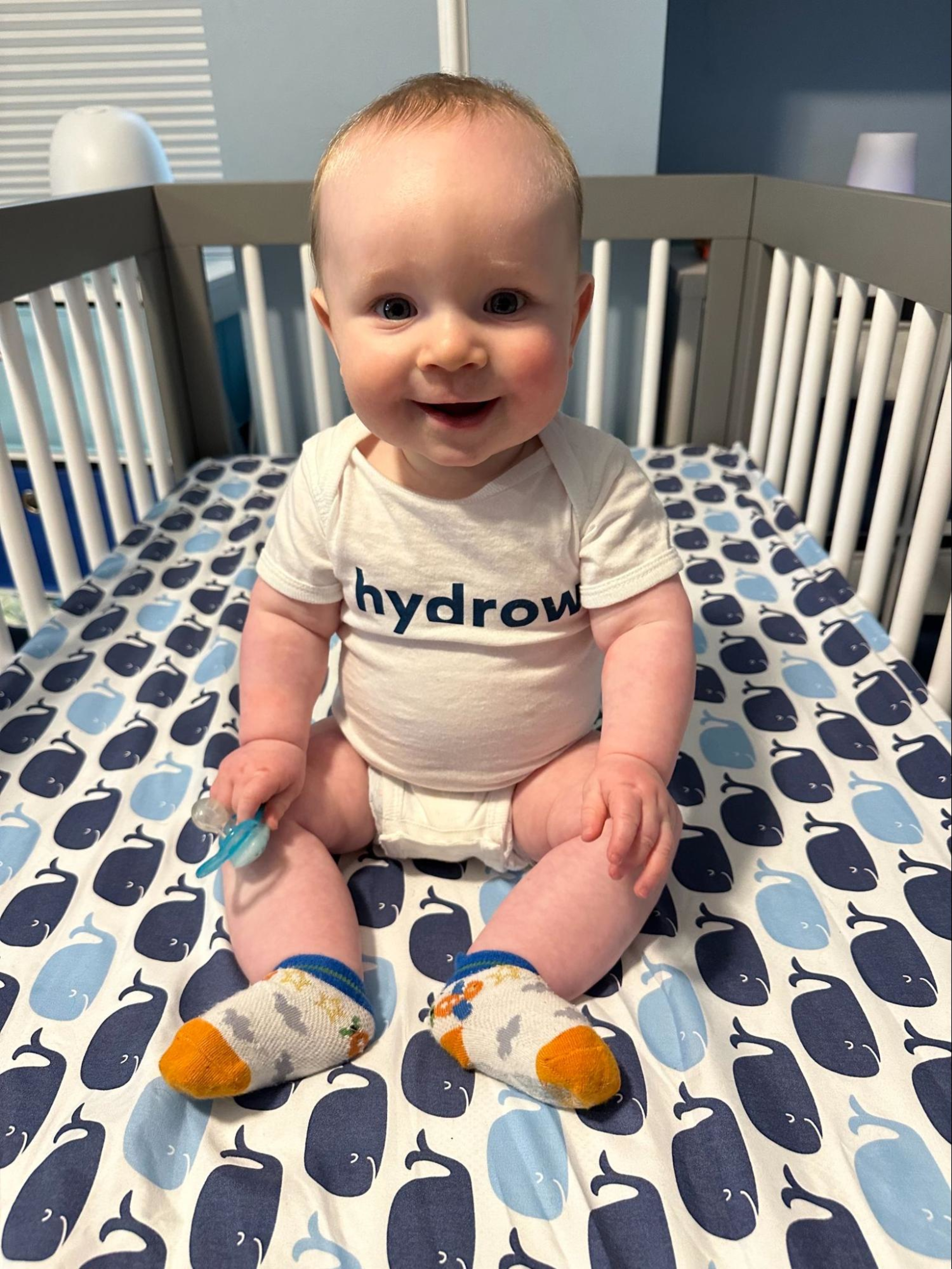 A baby sits in his crib wearing a Hydrow onesie.