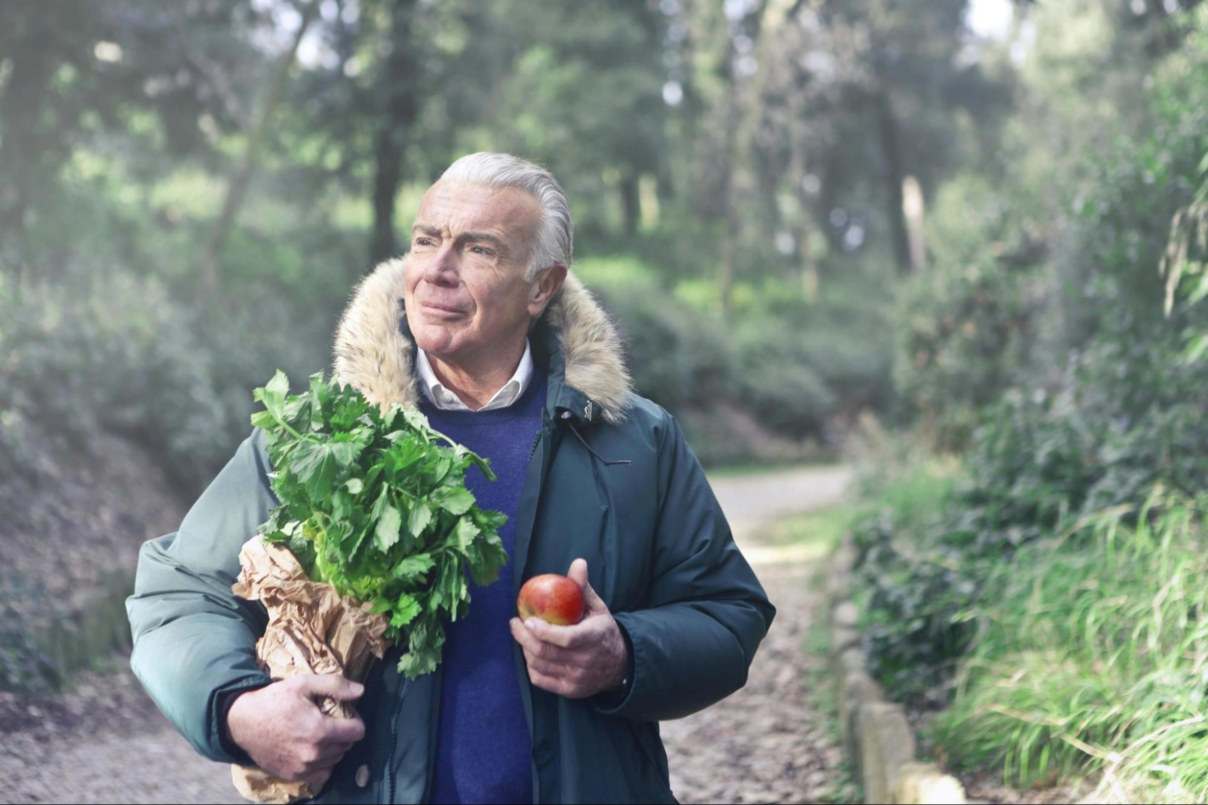 Older man walks with a bag of greens and an apple in his hand.