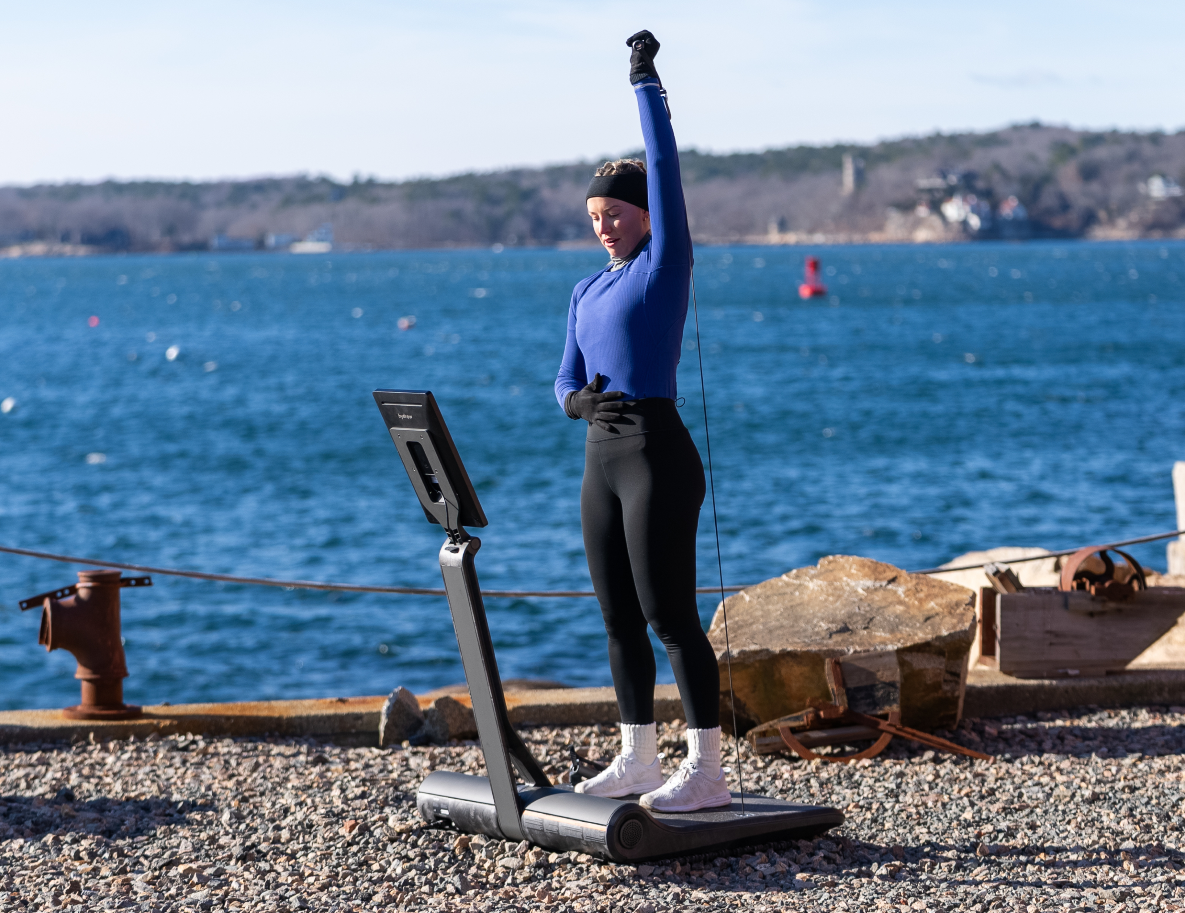 Hydrow Athlete Mac Evans does a single arm overhead press on a LYQUID strength machine outdoors.