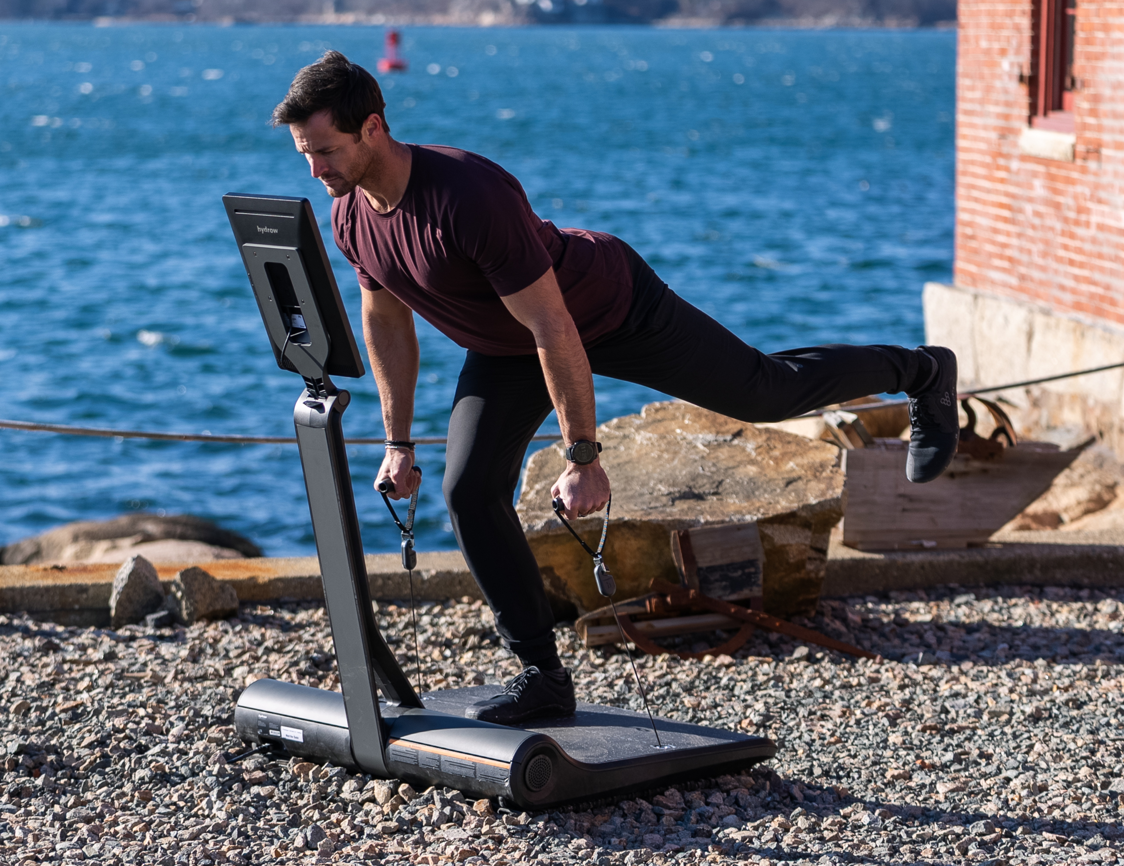 Hydrow Athlete Lance Parker does a single leg Romanian deadlift on a LYQUID strength machine outdoors.