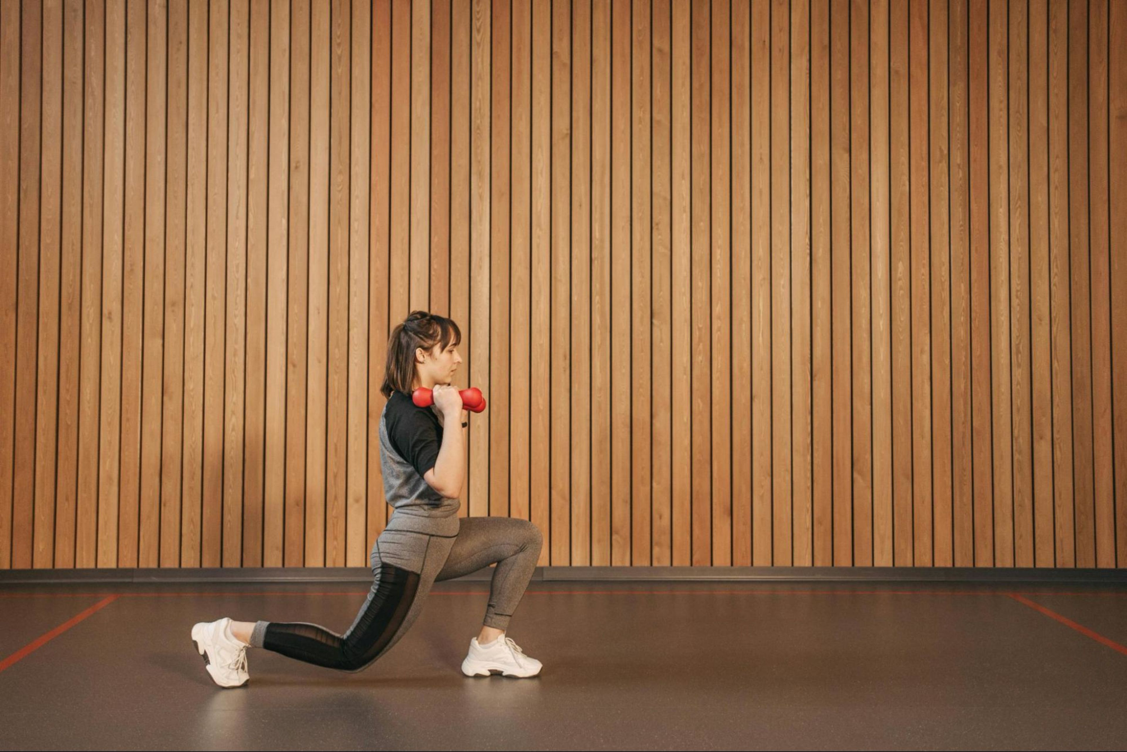 Woman does an easy leg exercise by lunging and holding a dumbbell in the gym.