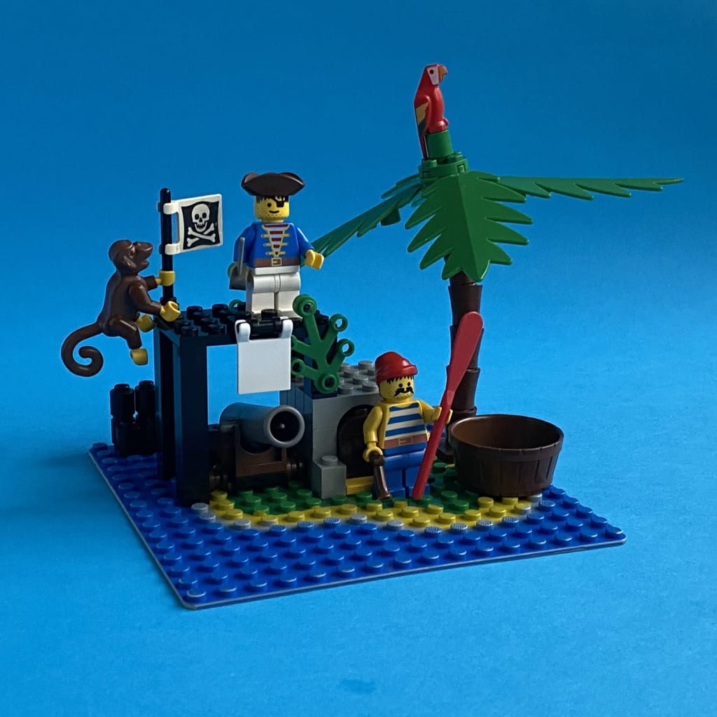 Small LEGO island with a palm tree and a ramshackle structure hiding a cannon and a treasure chest is inhabited by two pirates, a monkey and a parrot