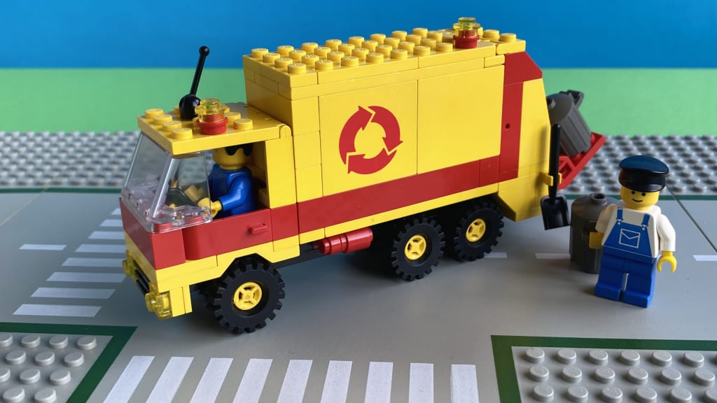 The finished Refuse Collection Truck from the side with the driver inside the cabin and the other minifigure carrying a bin next to the truck