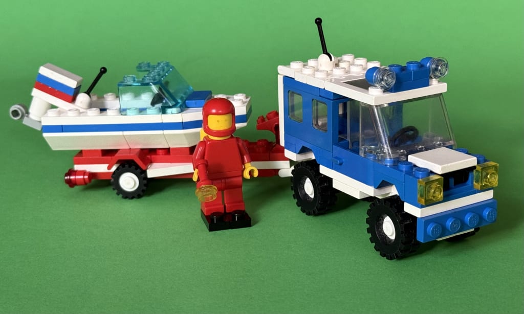 The minifigure wearing diving equipment standing in front of the vehicle and the attached trailer carrying the speedboat