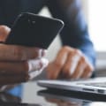 Person holds a cell phone while sitting in front of a laptop and calculator.