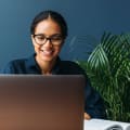 Woman in glasses uses a laptop on a desk with a notebook.