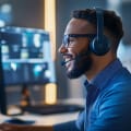A smiling man wearing headphones and a headset happily works in front of two computer monitors.