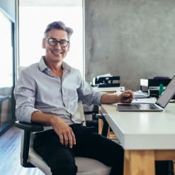 Smiling business person sits in front of a laptop with a writing implement.