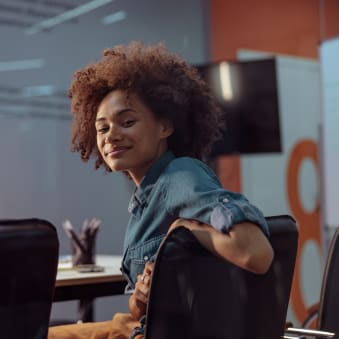 Person sits in conference room, turned around in seat and smiles at camera.