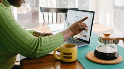 Person uses laptop at breakfast table with pot of coffee and coffee mug nearby.