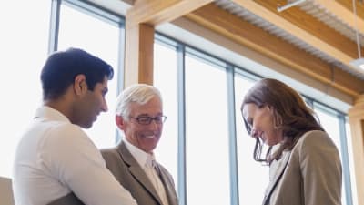 Three people smile and talk in an office. Two are holding clipboards. 