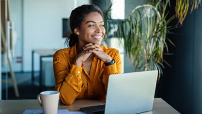 Person smiles and sits in front of a laptop computer and coffee mug.