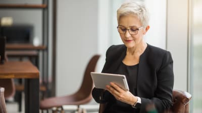 Person in dark suit sits in a wooden chair, working on a tablet computer.