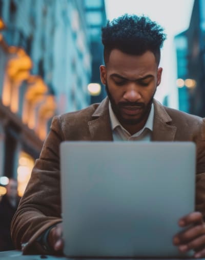 Focused young African American businessman using laptop on a busy city street at night.