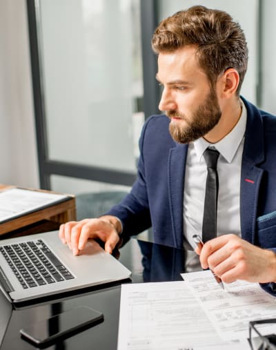 Person with a pen in hand sits and looks at a laptop in an office setting. There is paperwork on the table.