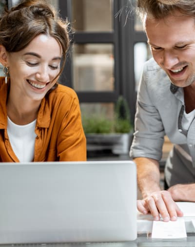 Two people lean over a table and look at a laptop. There are papers, a pair of glasses and a cup on the table.