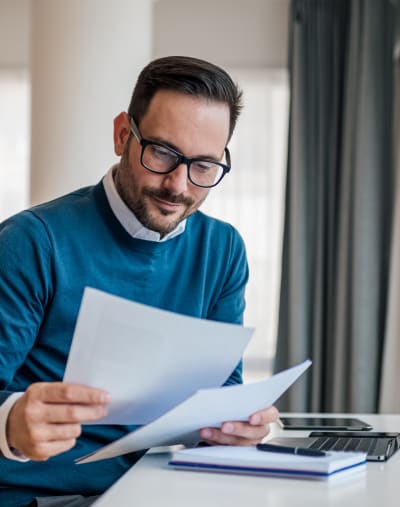 Person in glasses looks over paperwork. There is a notebook, laptop and mobile phone on the table.