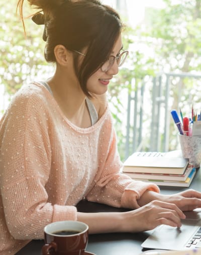 Person in glasses sits and types on a laptop. There is a cup of coffee and writing supplies on the table.