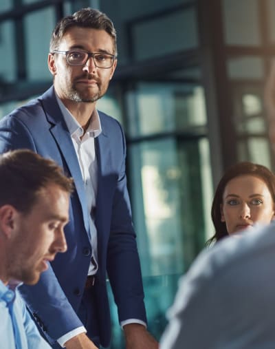 A group of people talk in an office. One person with glasses is standing as the others sit at a table. 