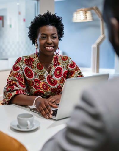 A person sits at a table with a laptop and a coffee cup and talks to a colleague.