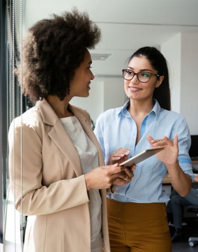 Two people interact in an office setting. One person is holding a tablet and the other has glasses.