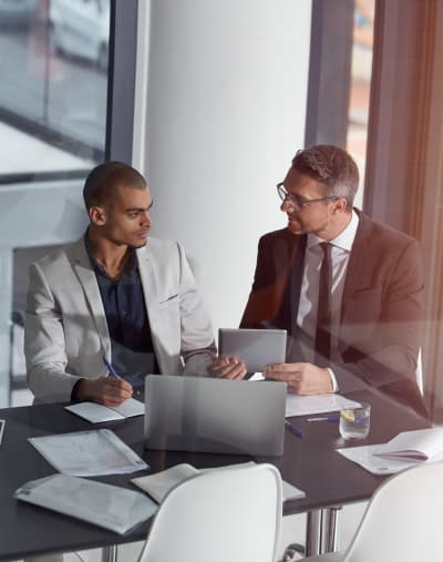 Two people sit and talk at an office table. One person shares information on a tablet.