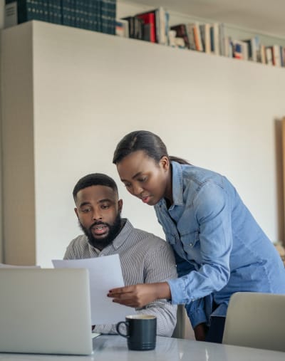 Two people look over paperwork. One is standing and leaning over the seated person’s shoulder.