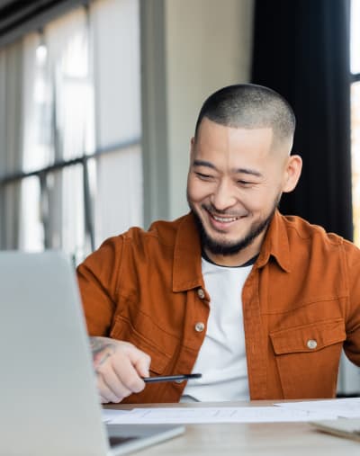 A person with a pen in hand smiles and looks at a laptop.