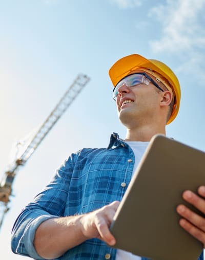 Construction worker in the field, wearing a hardhat and safety glasses while holding a tablet. 