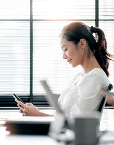 Person in casual business attire sits in an office setting and looks at a handheld device. 